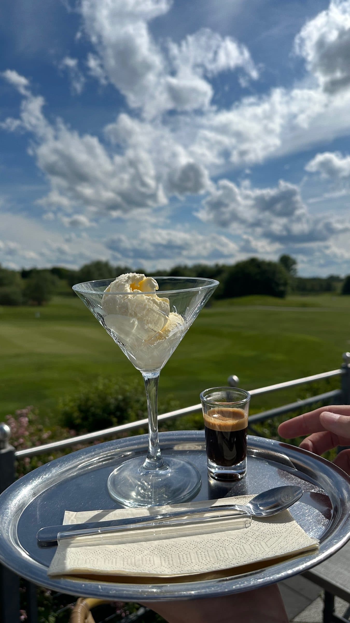 Vanilleeis im Glas und ein Espresso auf einem Tablett, dahinter Fairway und Sommerwolken