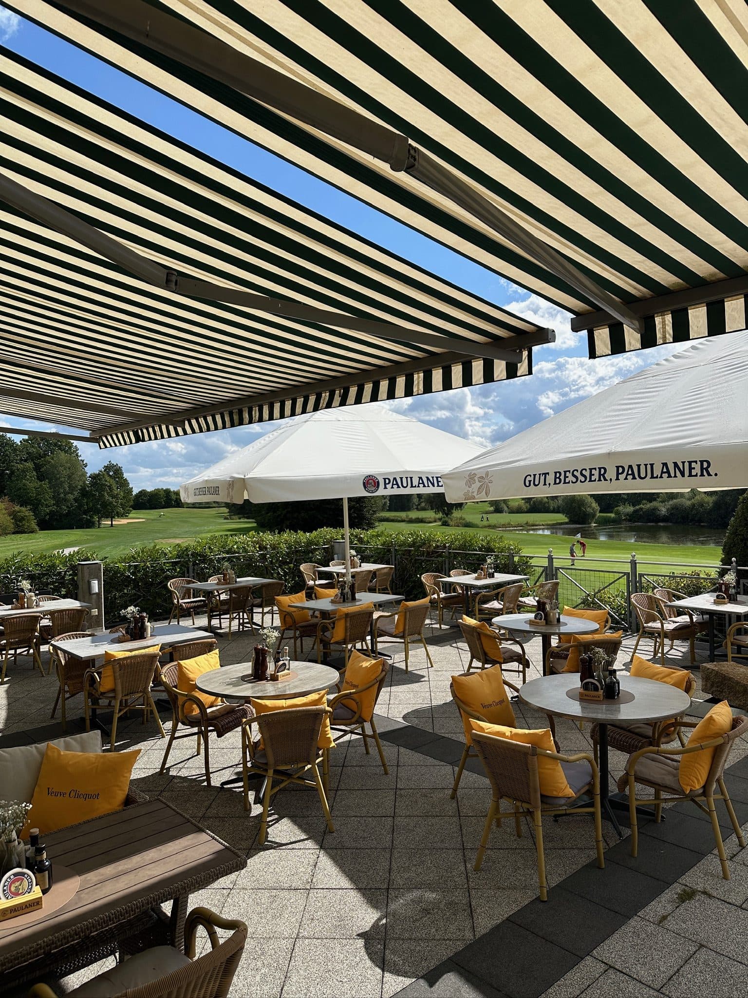 Eingedeckte Terrasse unter der gestreiften Markise, runde Tische mit gelben Kissen, dahinter Paulaner-Schirme und der Golfplatz
