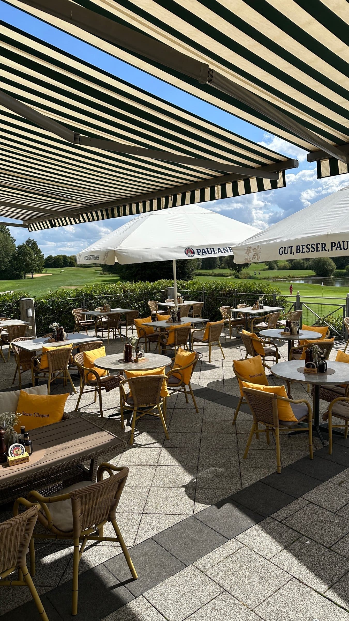 Blick unter der Markise auf die Terrasse mit Korbstühlen und Paulaner-Schirmen, dahinter die Fairways